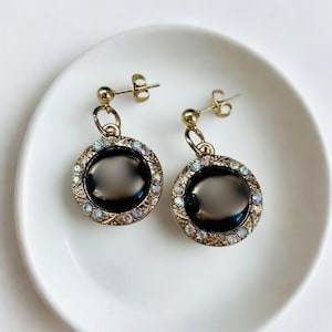 May include: A pair of gold-toned stud earrings. Each earring features a large, oval, black stone surrounded by a halo of iridescent crystals. The earrings are set on a white plate, showcasing their design and color.