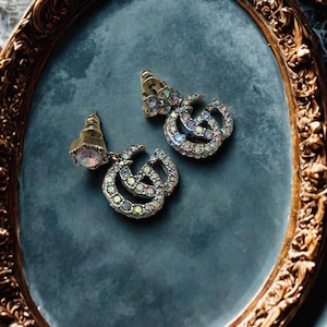 May include: A pair of silver-toned earrings with iridescent crystals. The earrings feature a double-G design and are set against a dark blue background within a decorative gold-framed oval.