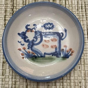 May include: A small, round ceramic bowl with a blue rim and a hand-painted cow design. The cow is depicted in blue with pink spots, surrounded by blue and orange floral accents. The bowl has a white interior.