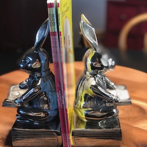 May include: Silver-colored bunny bookends holding children's books. The bookends are shaped like rabbits sitting and reading. The books have yellow, red, and white covers. The bookends are on a wooden tray.