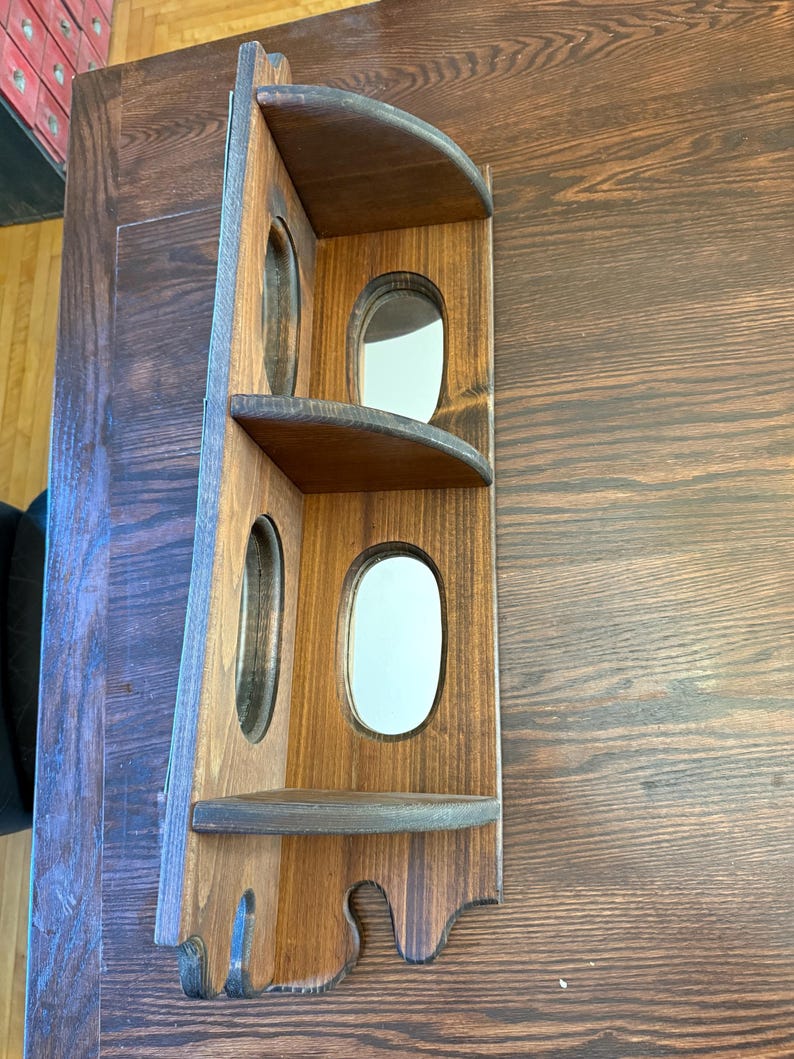 May include: A wooden corner shelf with a dark brown finish. It features two shelves and three oval openings. The shelf is designed to fit into a corner and has a decorative bottom edge. The wood grain is visible.