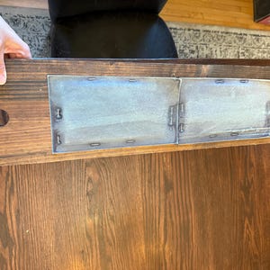 May include: A wooden table with a dark brown finish. The table has a rectangular shape with a decorative edge. Two metal panels are attached to the table's underside with small metal fasteners.