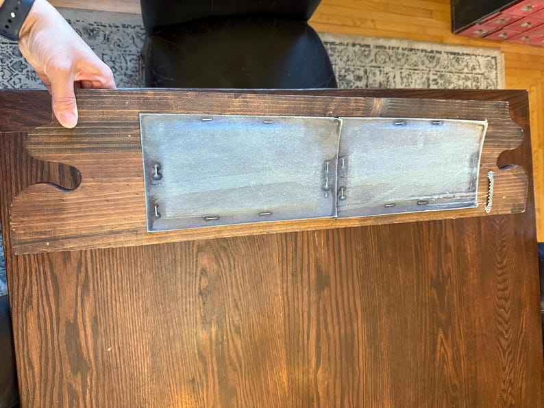 May include: A wooden table with a dark brown finish. The table has a rectangular shape with a decorative edge. Two metal panels are attached to the table's underside with small metal fasteners.