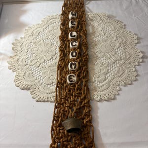 May include: Brown macrame wall hanging with a metal ring at the top. The macrame features six white ceramic beads with black letters spelling "BOO". A small bronze bell hangs at the bottom of the macrame.