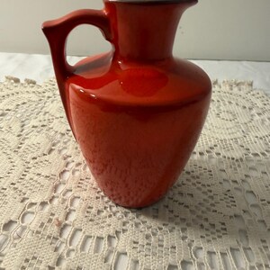 May include: A bright orange ceramic pitcher with a glossy finish, featuring a rounded body, a flared rim, and a single curved handle. The interior rim and the base of the handle show a dark brown tone, contrasting with the vibrant orange glaze.