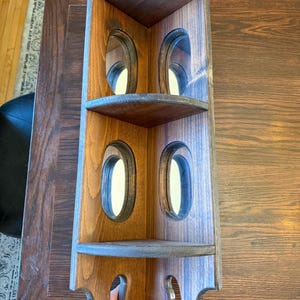 May include: A wooden corner shelf with three shelves and oval cutouts. The shelf is stained in shades of brown and grey, with decorative edges. The shelf is designed to fit into a corner and is suitable for displaying items.