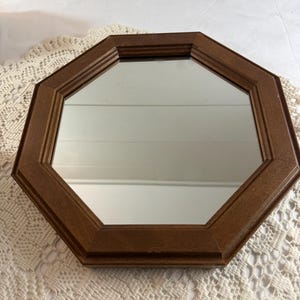 May include: An octagonal mirror with a dark brown wooden frame. The mirror's reflective surface is surrounded by the geometric frame, creating a decorative accent. The frame has a layered design, adding depth to the piece. The mirror is likely for wall decor.