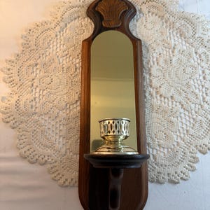 May include: A vintage wall-mounted candle holder with a wooden frame and a brass candle cup. The frame has a dark brown finish with a decorative shell carving at the top. The candle holder includes a small shelf and a brass-colored mirror.