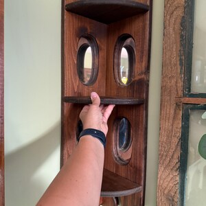 May include: A dark stained wooden corner shelf with three shelves and oval cutouts. The shelf is mounted on a light green wall. The shelf is designed to fit into a corner and has a decorative top edge.