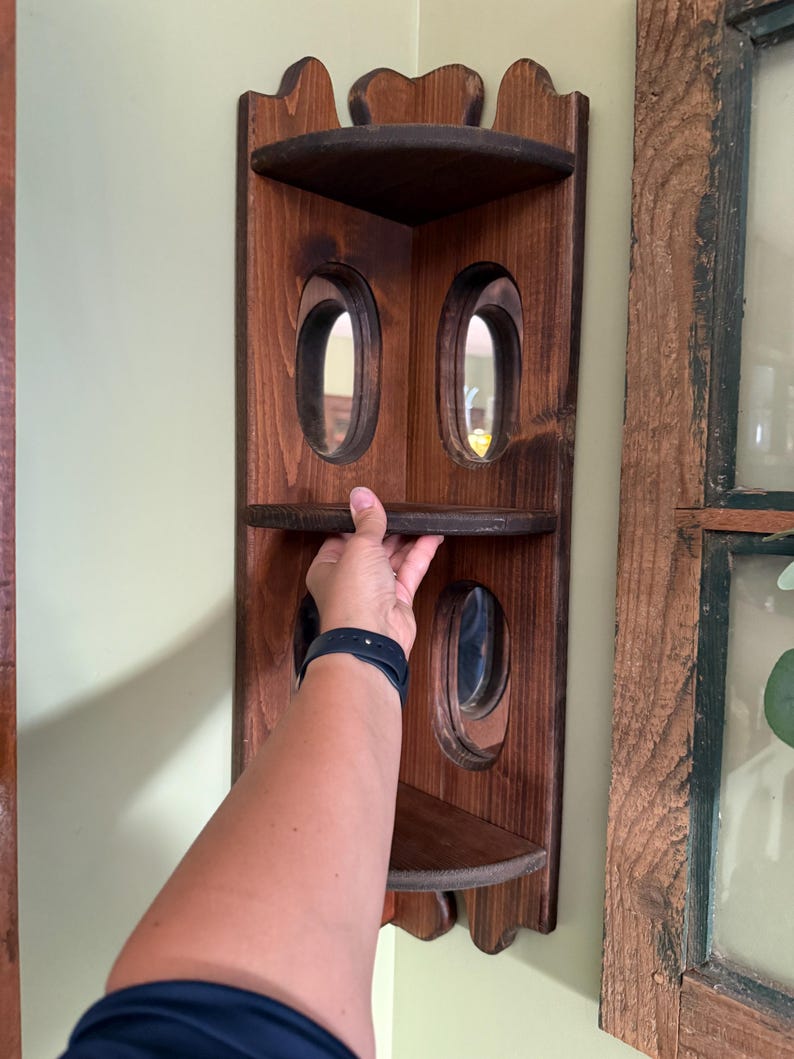 May include: A dark stained wooden corner shelf with three shelves and oval cutouts. The shelf is mounted on a light green wall. The shelf is designed to fit into a corner and has a decorative top edge.