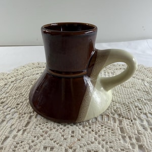 May include: A brown and cream-colored ceramic mug with a unique, flared shape. The mug features a dark brown upper section and a lighter cream-colored handle and lower portion. The handle is a curved, rounded shape.