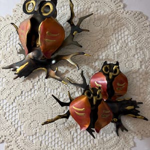 Könnte beinhalten: Zwei Metall-Eulen-Skulpturen auf Ästen. Die Eulen haben schwarze und goldene Details mit kupferfarbenen Flügeln. Die Äste sind dunkelbraun mit goldenen Akzenten. Die Skulpturen sind vor einem cremefarbenen, spitzenartigen Hintergrund platziert.