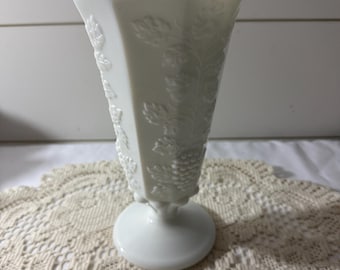 May include: A white, vintage vase with a flared, scalloped rim. The vase has embossed grape and leaf designs on its sides. It has a pedestal base and is made of opaque glass. The vase is a decorative item, suitable for displaying flowers or as a standalone piece.