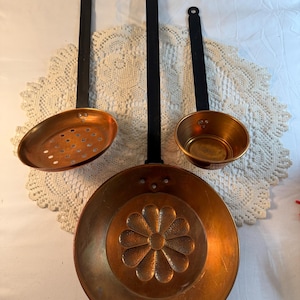 May include: A collection of antique copper kitchenware, including a large pan with a floral design, a slotted spoon, and a small ladle. Each piece has a copper bowl and a black handle.