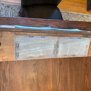May include: A dark brown wooden desk with a decorative edge and two metal panels. The desk has a rectangular shape and is positioned on a wooden floor. The metal panels are attached with small metal staples.