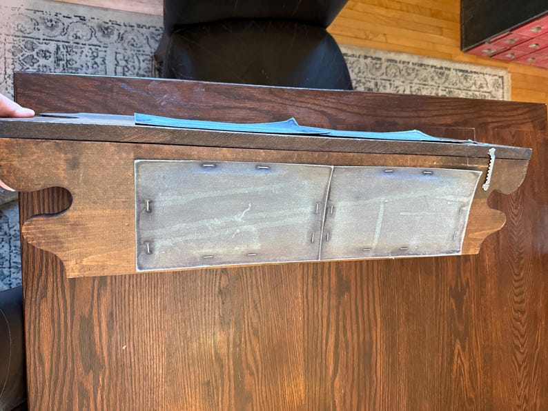 May include: A dark brown wooden desk with a decorative edge and two metal panels. The desk has a rectangular shape and is positioned on a wooden floor. The metal panels are attached with small metal staples.