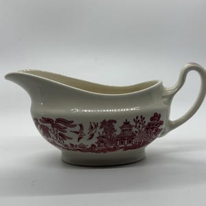 May include: A white ceramic gravy boat with a pink willow pattern design. The boat has a rounded body and a handle on the right side.
