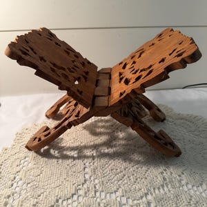 May include: A brown wooden book stand featuring intricate cut-out patterns on both panels. The stand is constructed from two interlocking pieces that form an X-shape, designed to hold a book open with its carved, decorative edges and support legs.