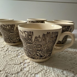 Könnte beinhalten: Sechs cremefarbene Keramik-Teetassen mit einem braunen Transferdruck, der eine historische Stadtszene zeigt. Jede Tasse hat einen gebogenen Henkel und einen breiten Rand. Die Tassen sind auf einer weißen Spitzendecke angeordnet.