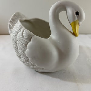 Puede incluir: Una maceta de cisne de cerámica blanca con pico amarillo y ojos negros. El cisne tiene un diseño detallado de plumas en la espalda y las alas, y un área ahuecada para plantar flores o guardar objetos pequeños.