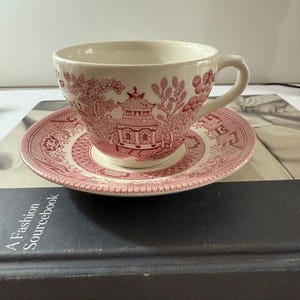 Puede incluir: Una taza de té y un platillo rosa y blanco con un patrón floral. El patrón presenta una pagoda y árboles. La taza de té y el platillo están sobre un libro.