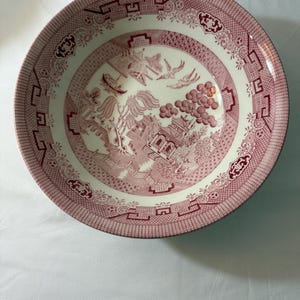 Op de afbeelding: Een ronde keramische kom met een rood en wit Willow patroon. De kom heeft een gedetailleerde landschapsweergave met pagodes, vogels en bloemelementen. De rand heeft een decoratieve rand. De kom wordt waarschijnlijk gebruikt voor het serveren van voedsel.