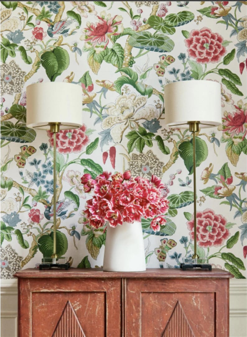 May include: Two white lamps with gold bases stand on a wooden dresser with a floral wallpaper background. A white vase with pink flowers sits on the dresser between the lamps.