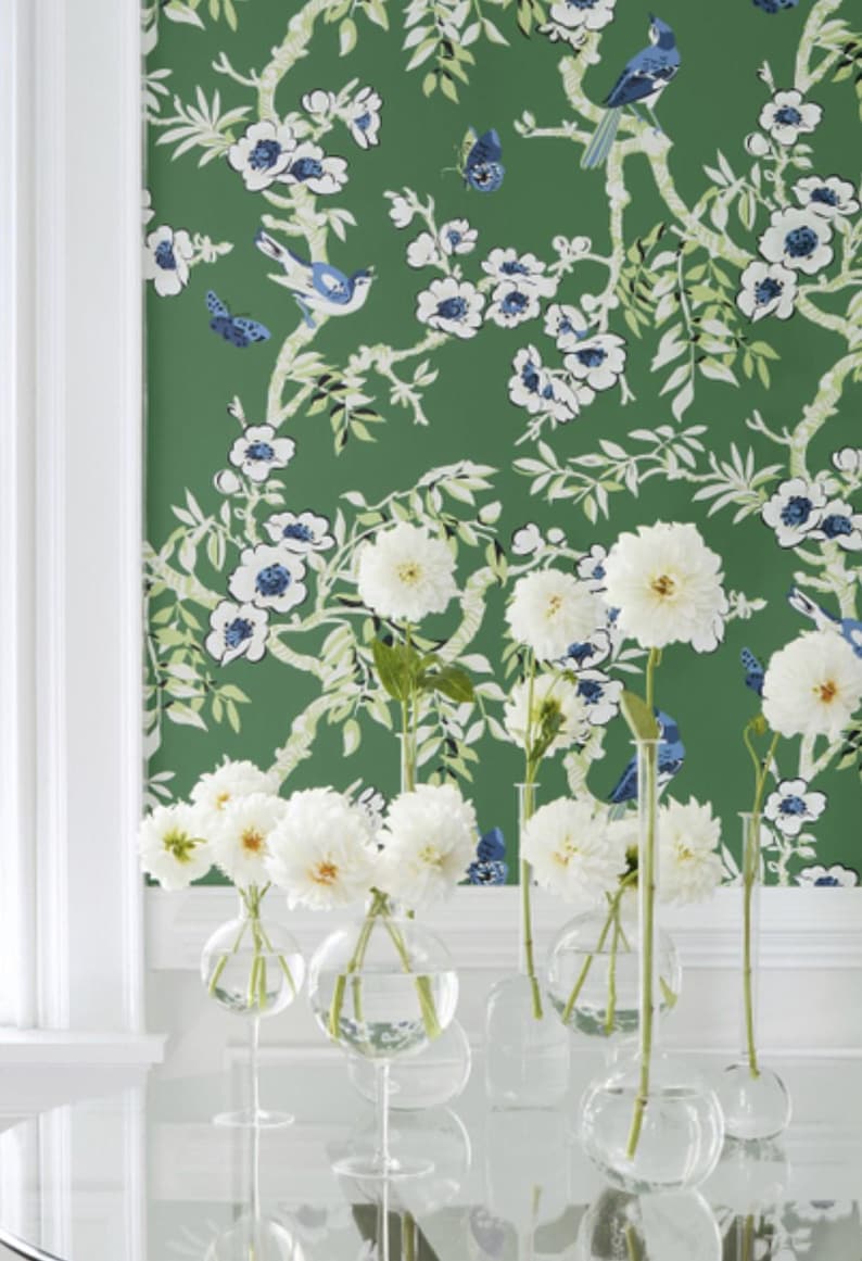 May include: White flowers in clear glass vases with round bulbs. The vases are on a white table with a green floral wallpaper in the background.