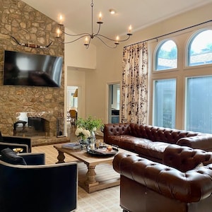 May include: A living room with a stone fireplace, a brown leather Chesterfield sofa, and two black armchairs. A wooden coffee table sits in front of the sofa, and floral curtains frame the large windows. A decorative chandelier hangs from the ceiling.