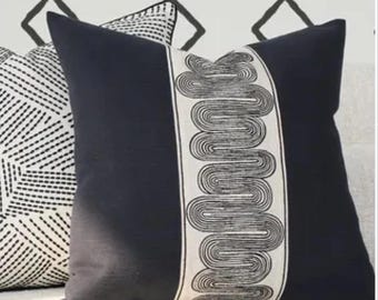 May include: A navy blue decorative pillow with a cream-colored vertical stripe featuring a black, wavy line design. Another pillow with a black and white geometric pattern is partially visible. The background shows a white wall with black diamond shapes.
