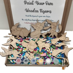 May include: A wooden craft kit featuring 10 unpainted wooden figures of Pokemon characters, including Eevee, Vaporeon, Jolteon, Flareon, and more. The kit also includes six small bottles of acrylic paint and two paintbrushes. The figures are in a cardboard box with colourful shredded paper.
