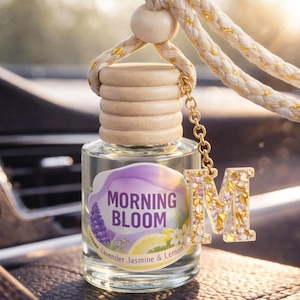 May include: A car air freshener in a clear glass bottle with a wooden cap. The label says "MORNING BLOOM" with lavender, jasmine, and lemon illustrations. A gold-coloured charm with the letter "M" hangs from the bottle.
