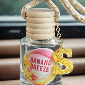 May include: Clear glass car air freshener bottle with a wooden cap and a gold-colored chain. The label reads "Banana Breeze" with a banana and candy graphic. A gold letter "S" charm hangs from the chain.