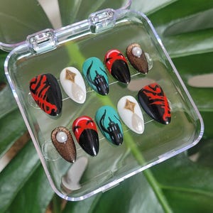 May include: A set of ten artificial nails in a clear plastic case. The nails feature various designs, including red and black patterns, teal cityscapes, and gold accents with pearls. The nails are oval-shaped and have a glossy finish.