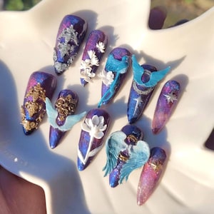 Puede incluir: Una colección de uñas artificiales largas en forma de almendra. Cada uña presenta una base iridiscente de color púrpura y azul oscuro con varios adornos, que incluyen filigranas plateadas, diseños florales blancos y pequeños acentos de alas de pájaros y ángeles azules. Las uñas están dispuestas sobre un plato blanco en forma de hoja.