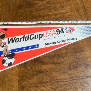 K&ouml;nnte beinhalten: Ein rot-wei&szlig;-blaues Wimpel mit einem Cartoon-Hund, der einen Fu&szlig;ball tritt. Der Wimpel tr&auml;gt die Aufschrift "World Cup USA 94" und "Making Soccer History".