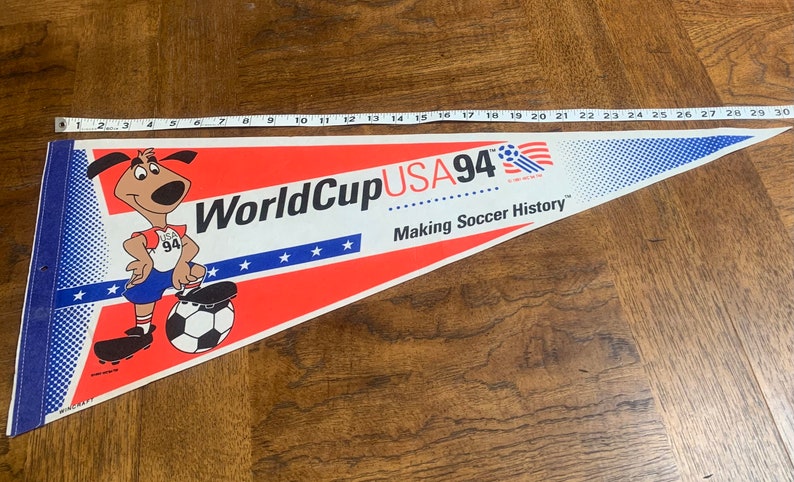K&ouml;nnte beinhalten: Ein rot-wei&szlig;-blaues Wimpel mit einem Cartoon-Hund, der einen Fu&szlig;ball tritt. Der Wimpel tr&auml;gt die Aufschrift "World Cup USA 94" und "Making Soccer History".