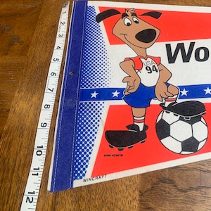 K&ouml;nnte beinhalten: Ein rot-wei&szlig;-blaues Wimpel mit einem Cartoon-Hund, der ein Fu&szlig;balltrikot mit dem Text "USA 94" tr&auml;gt. Der Hund h&auml;lt einen Fu&szlig;ball und steht auf einem Feld mit Sternen. Der Wimpel wird von Wincraft hergestellt.