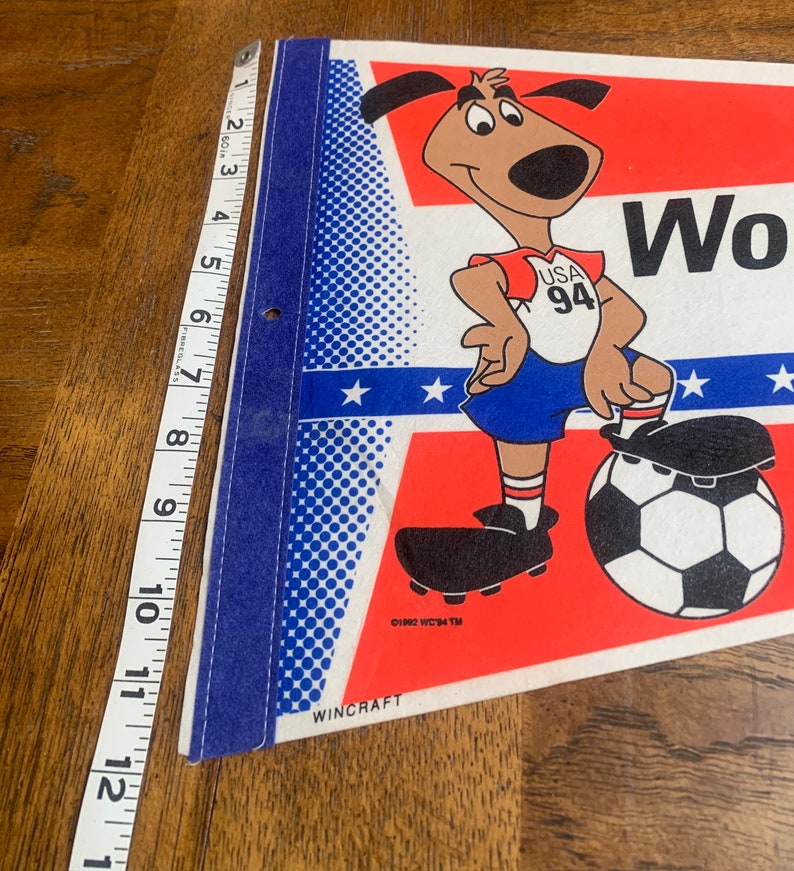 K&ouml;nnte beinhalten: Ein rot-wei&szlig;-blaues Wimpel mit einem Cartoon-Hund, der ein Fu&szlig;balltrikot mit dem Text "USA 94" tr&auml;gt. Der Hund h&auml;lt einen Fu&szlig;ball und steht auf einem Feld mit Sternen. Der Wimpel wird von Wincraft hergestellt.