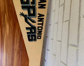 May include: A vintage San Antonio Spurs pennant with the team's logo and name in black and white on a tan background.