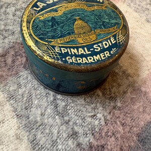 May include: Vintage round tin with a blue and gold design. The lid features text in French, including "LA SEVE DE PIN" and "BONBONS". The tin has a detailed illustration of a landscape with a beehive. The tin is likely a container for candies.
