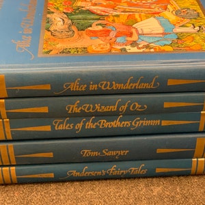 May include: A stack of classic blue hardcover books with gold lettering. Titles include "Alice in Wonderland," "The Wizard of Oz," and "Tom Sawyer." The books have gold accents and are stacked on top of each other.