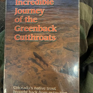 Puede incluir: Una portada de libro con el título "Incredible Journey of the Greenback Cutthroats" y el subtítulo "Colorado's native trout brought back from extinction". La portada presenta una foto de un lecho de arroyo con una trucha nadando en el agua.