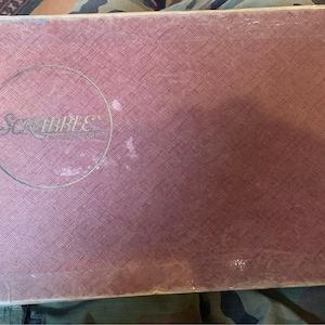 May include: A vintage Scrabble game box with a pink fabric cover and the word "Scrabble" printed in a circle on the front.