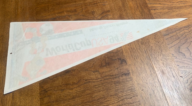K&ouml;nnte beinhalten: Ein wei&szlig;er Wimpel mit einem roten und blauen Design. Der Wimpel zeigt einen Fu&szlig;ballspieler, der einen Ball tritt, und den Text "World Soccer World" und "Mountaineers".