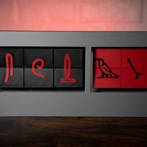 May include: A set of six black and red wooden blocks with hieroglyphic symbols carved into them. The blocks are arranged in a row in a gray frame.