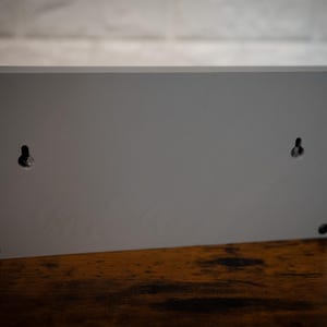 May include: The back of a gray rectangular sign with two small holes for hanging. The sign is made of wood and has a smooth surface.