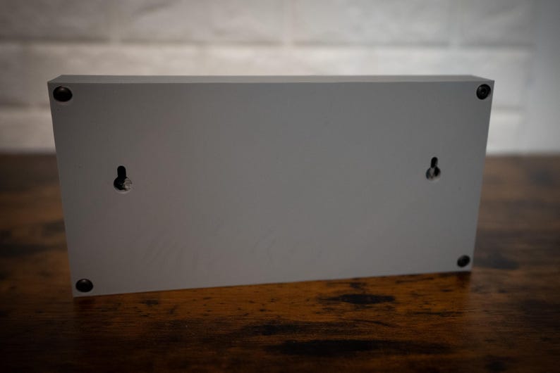 May include: The back of a gray rectangular sign with two small holes for hanging. The sign is made of wood and has a smooth surface.