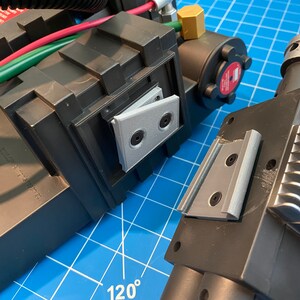 May include: Close-up of a black and silver toy proton pack with a gray plastic hinge. The pack has a textured surface and a silver metal grill.