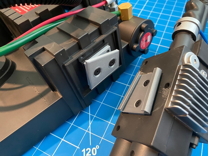 May include: Close-up of a black and silver toy proton pack with a gray plastic hinge. The pack has a textured surface and a silver metal grill.